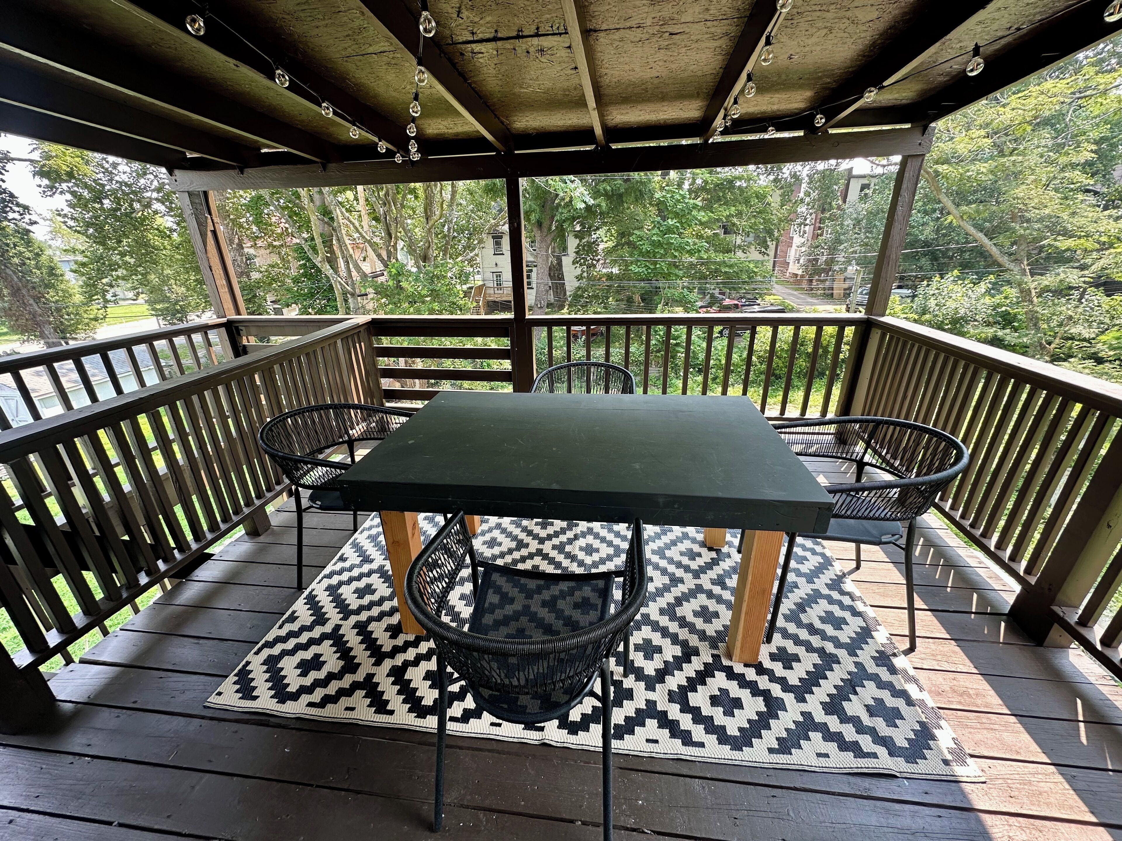 En täckt uteplats med ett bord och stolar, omgiven av ett trästaket, med utsikt över ett bostadsområde med träd och hus.