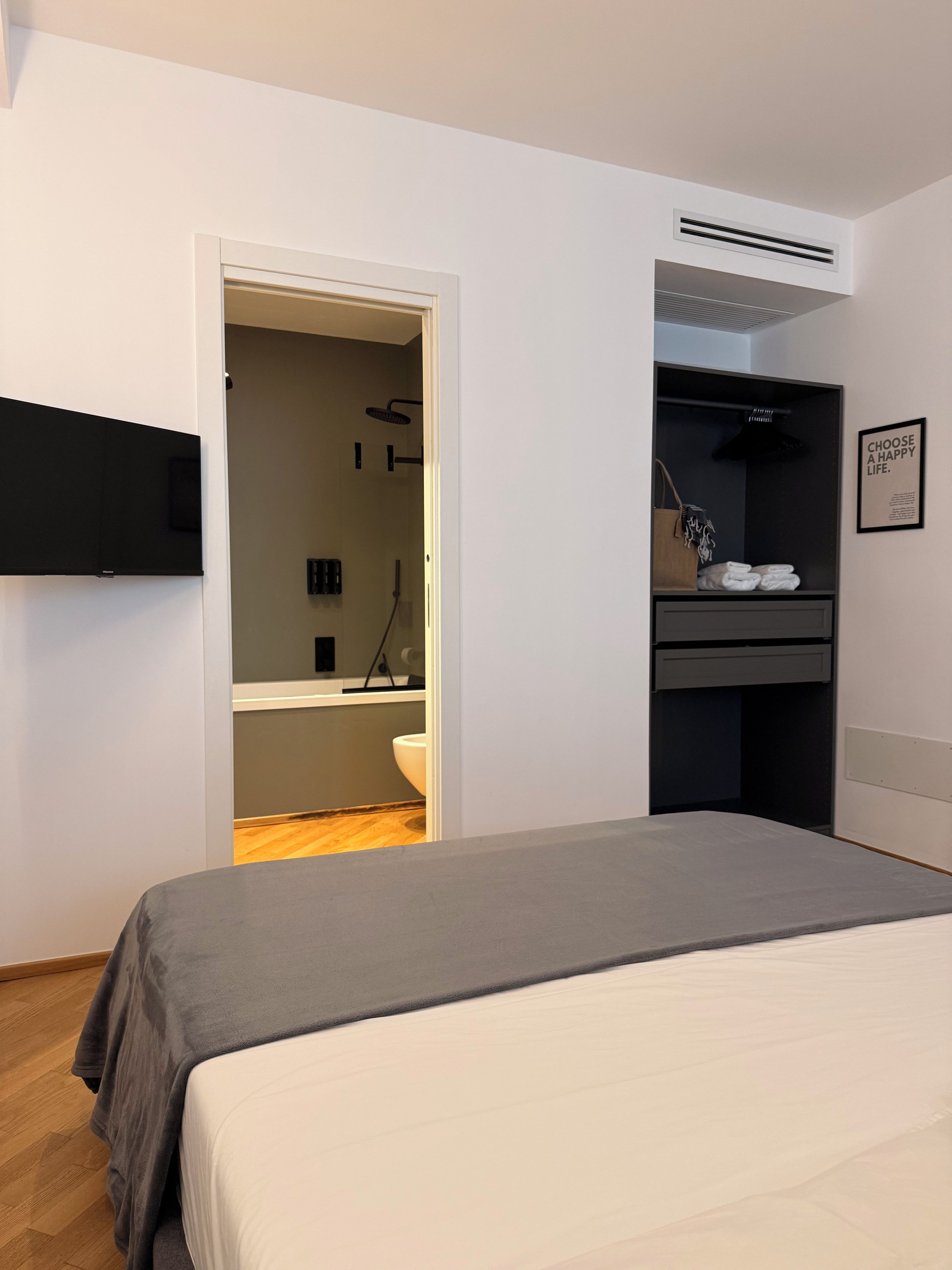 Une chambre à coucher avec un lit, un téléviseur fixé au mur, un dressing et une salle de bain visible à travers une porte ouverte.