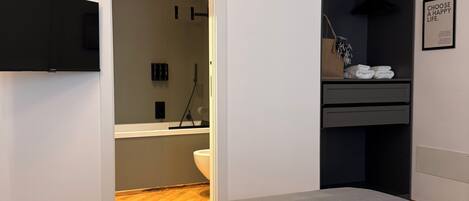Une chambre à coucher avec un lit, un téléviseur fixé au mur, un dressing et une salle de bain visible à travers une porte ouverte.