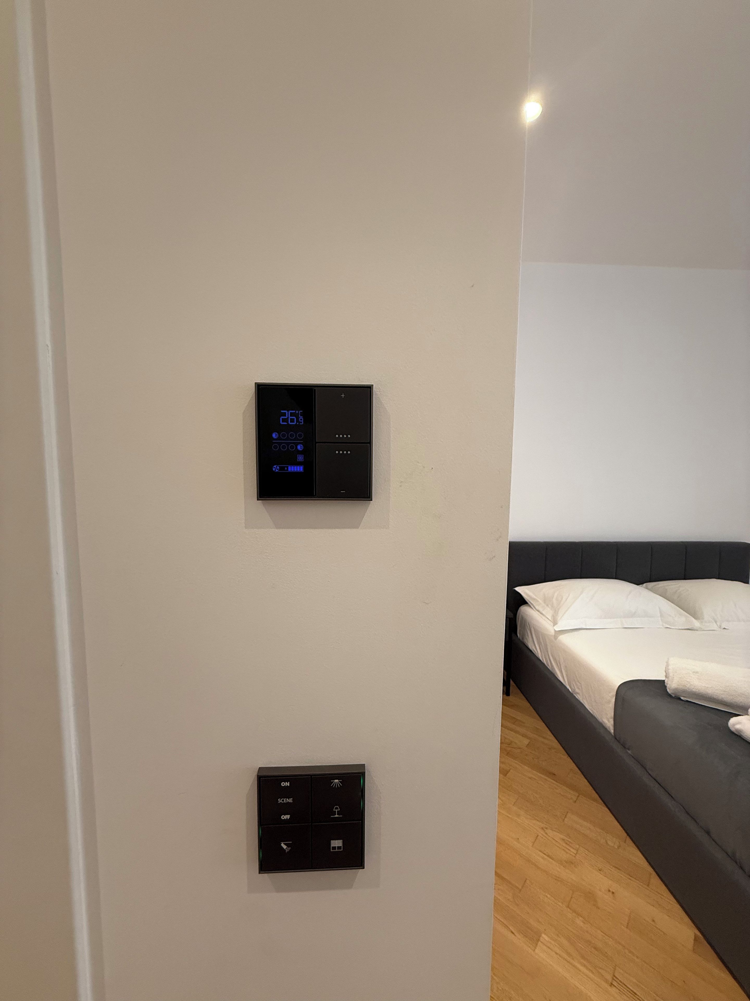 A room with a bed, a wall-mounted thermostat, and a control panel with lighting options.
