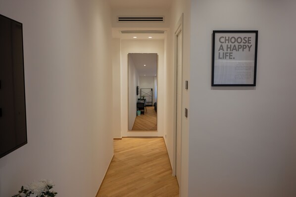 A hallway with a framed inspirational quote on the wall, a wooden floor, and a mirror reflecting a room with a bed and a desk.