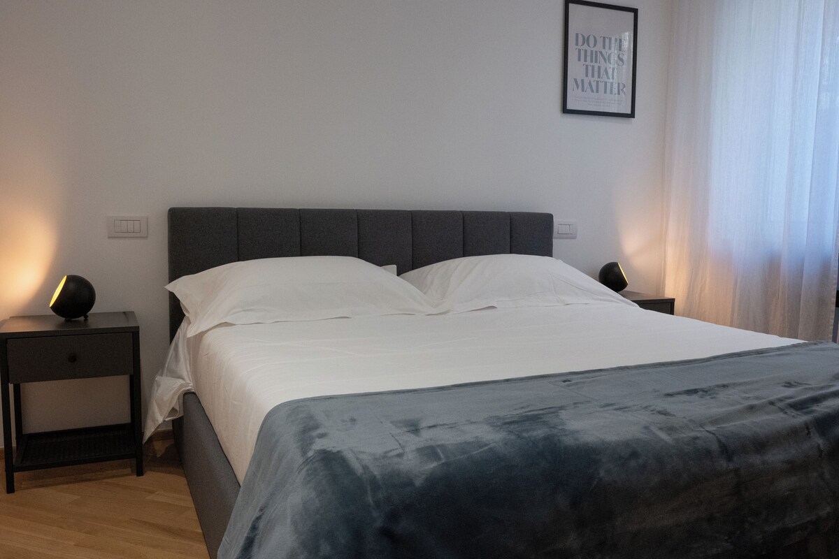 Una cama bien hecha con sábanas blancas y cabecera gris, una mesita de noche con lámpara, y una cita enmarcada en la pared.