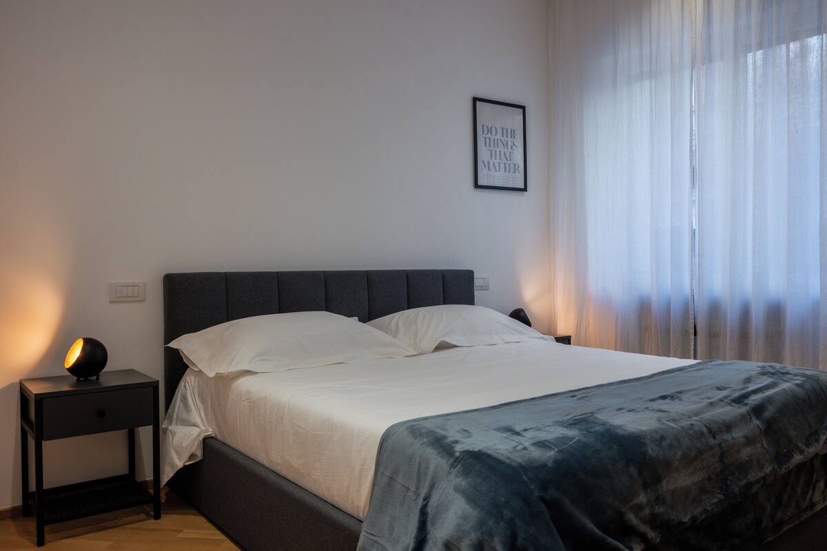 Un dormitorio con cama, una mesita de noche con lámpara, una cita enmarcadas en la pared y una ventana con cortinas de gasa.