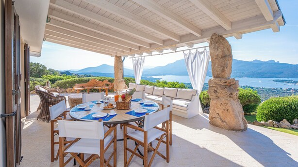 An outdoor patio with a round table set for dining, a sofa, and a view of the sea and mountains.