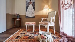 A room with a patterned rug, a white chair, a small table with a vase, and a painting of a woman in a dress on the wall. - CR BnB Rozzano (Rozzano)
