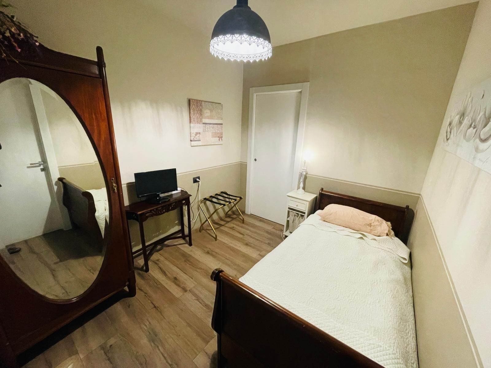 Een slaapkamer met een bed, een televisie op een houten standaard, een bureau met een stoel, een hanglamp en een garderobe.