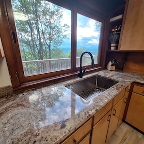 A kitchen with a granite countertop, a stainless steel sink, wooden cabinets, and a window with a view of trees and the sky.
