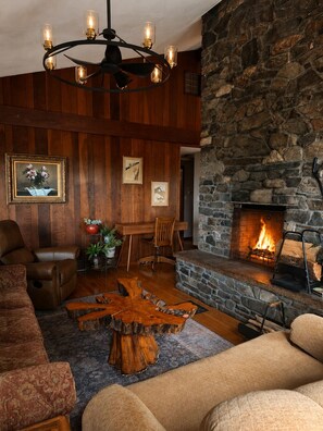 A cozy living room with a stone fireplace, wooden paneling, a chandelier, and a dining area.