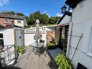 A wooden deck with a glass table and chairs, surrounded by potted plants and a hanging light fixture.