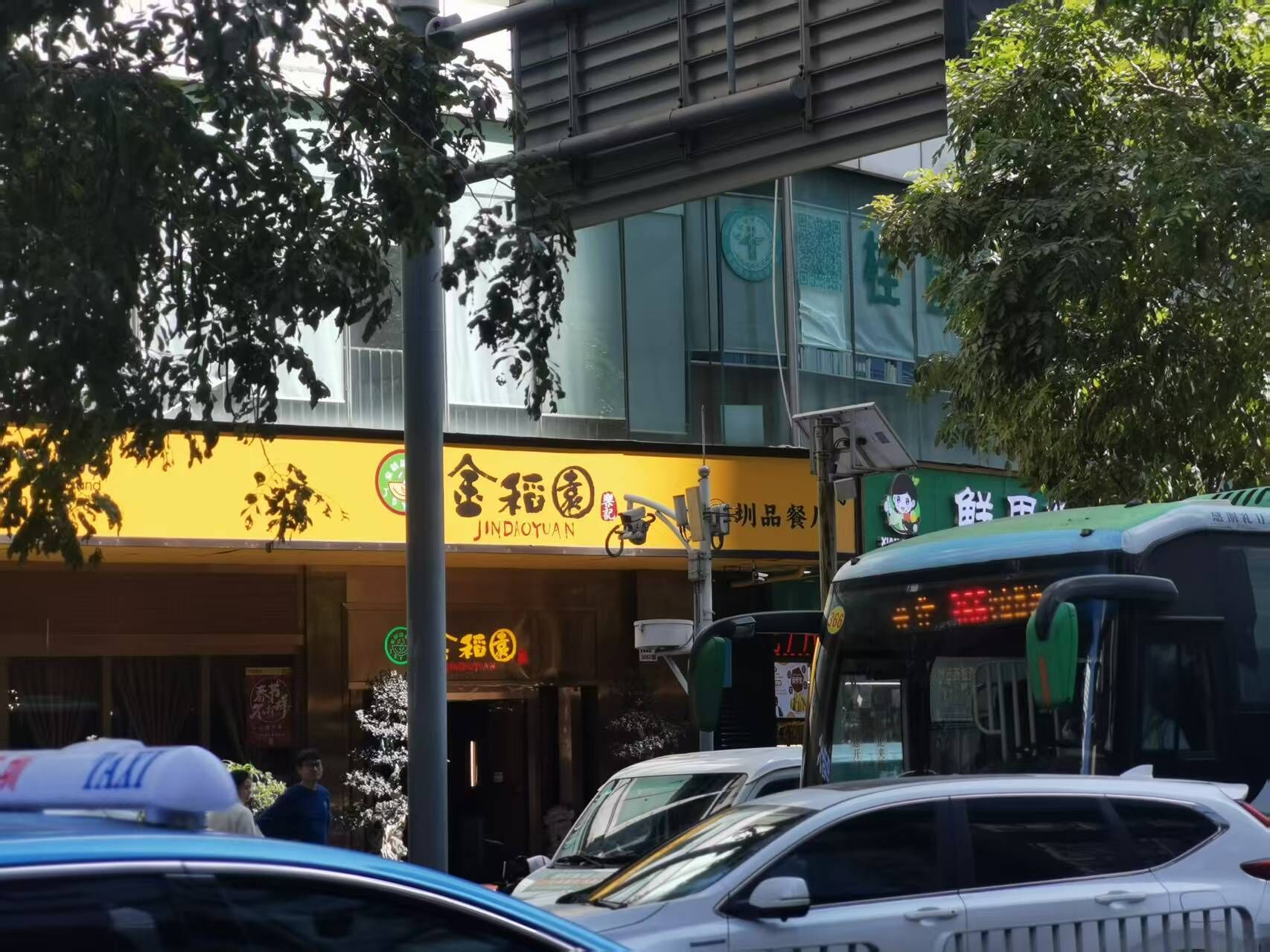 Cena de rua com um edifício que possui um restaurante chamado 'Jindaoyuan' e um letreiro amarelo. Há carros estacionados em frente e um ônibus visível à direita.