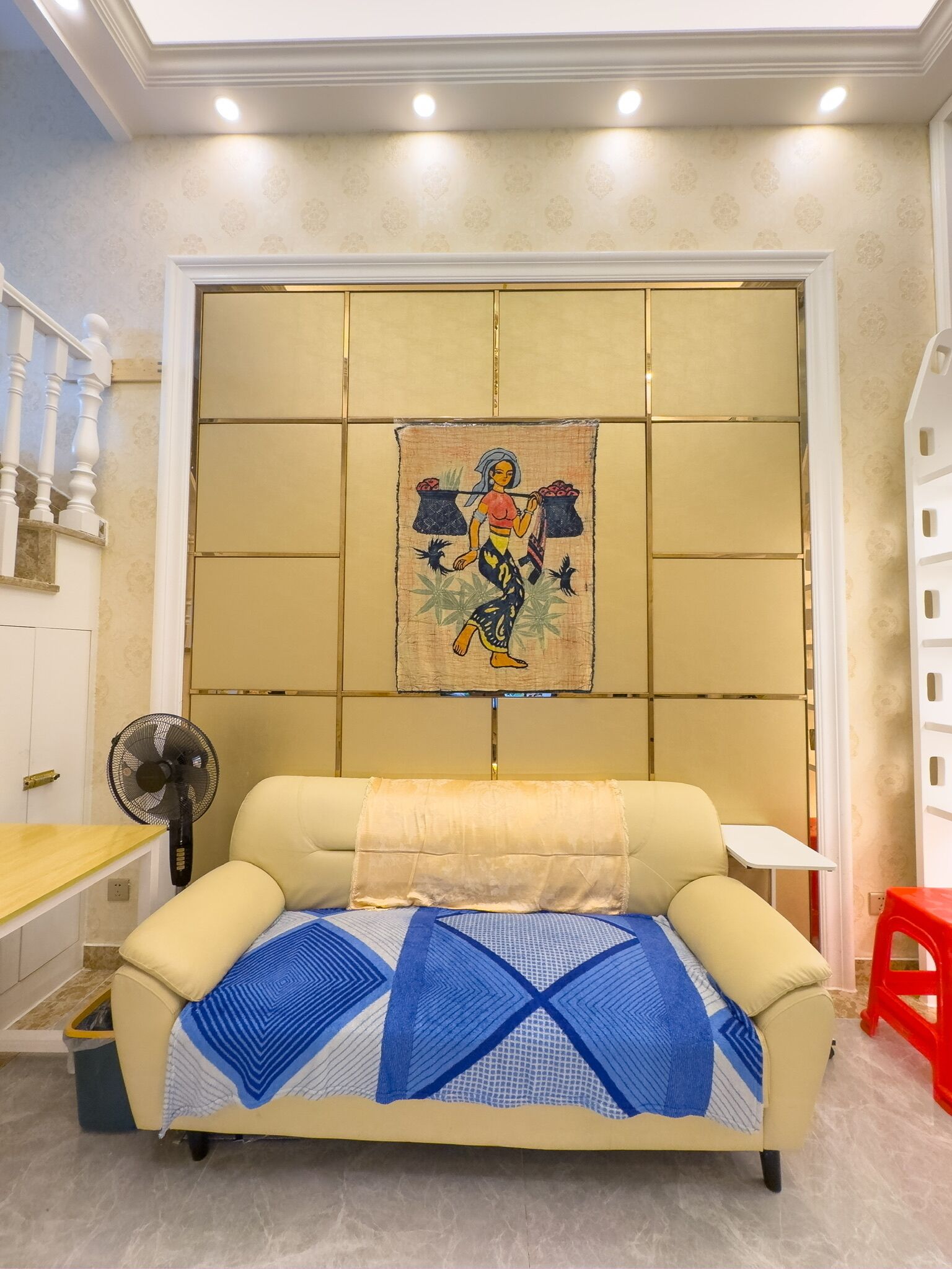 Un salon avec un canapé beige, un plaid à motifs, une tapisserie murale, un ventilateur sur pied et un petit tabouret rouge.