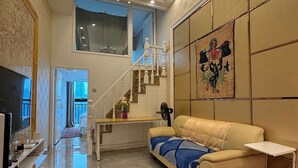 A spacious living room with a high ceiling, a chandelier, a sofa, a television, and a staircase leading to an upper level. - New CBD high-rise 3-brm LOFT, Huaxun Center, Luohu (Shenzhen)