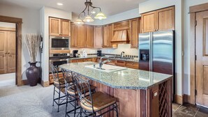 A modern kitchen with wooden cabinets, a stainless steel refrigerator, a granite countertop, and a bar area with stools. - Luxurious Ski-In/Ski-Out Condominium in the Heart of Mountain Paradise (Keystone)