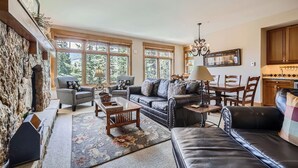 A spacious living room with a stone fireplace, leather sofas, a dining area with wooden chairs, and large windows offering a view of greenery. - Luxurious Ski-In/Ski-Out Condominium in the Heart of Mountain Paradise (Keystone)