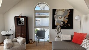Un salon de style industriel avec un haut plafond, une grande fenêtre, un canapé gris, un fauteuil en forme de poire, une petite table avec des haut-parleurs et un grand tableau abstrait.