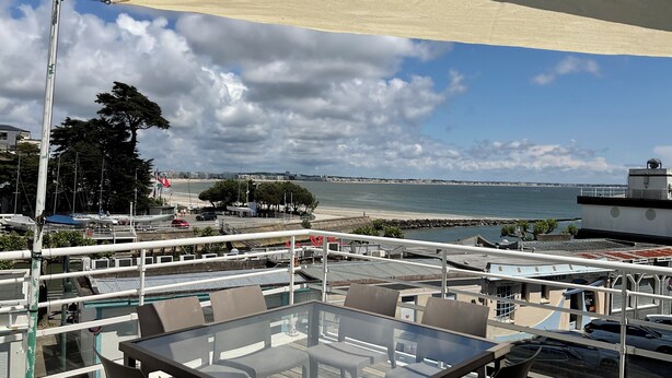 Une terrasse dotée de tables et de chaises à plateaux en verre, offrant une vue sur une marina avec des bateaux et une plage.