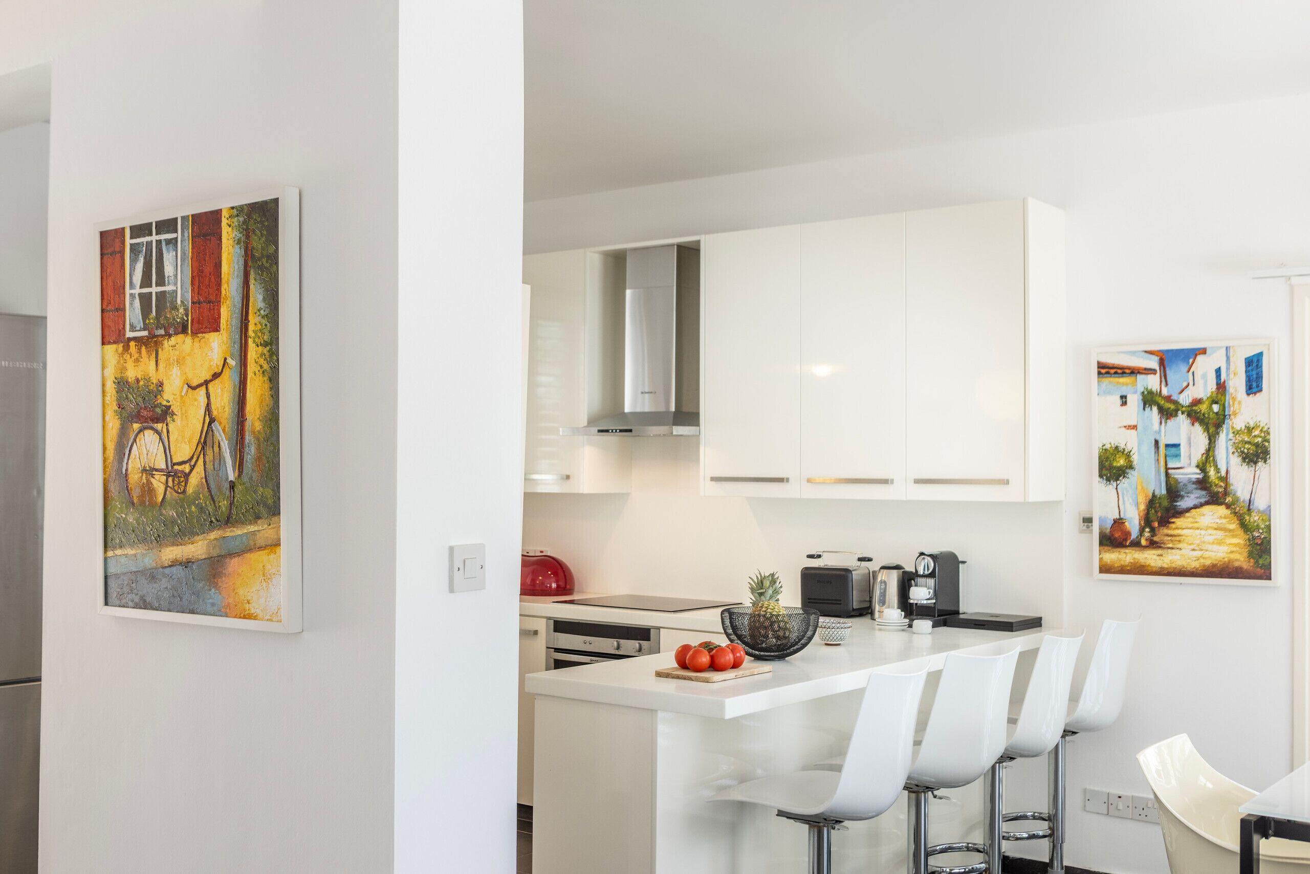 A modern kitchen with white cabinets, a central island with bar stools, a red kettle, and a pineapple on the counter.