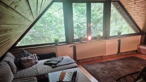 Une pièce mansardée chaleureuse avec un lit en mezzanine, une petite table avec un jeu d’échecs, un fauteuil en osier et une vue sur la verdure grâce à de grandes fenêtres.
