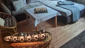 Une pièce chaleureuse avec un plafond en bois, un canapé avec des coussins, une petite table sur laquelle est posé un jeu d’échecs et un fauteuil en osier.