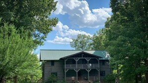 Un chalet rustique en rondins de bois, doté d’une véranda et entouré d’une végétation luxuriante et d’arbres.