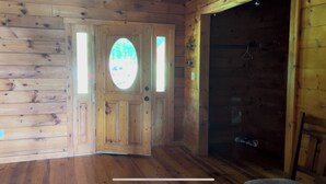 L’intérieur d’un chalet en bois, avec une porte donnant sur une véranda, un espace douche et une fenêtre offrant une vue sur la verdure.
