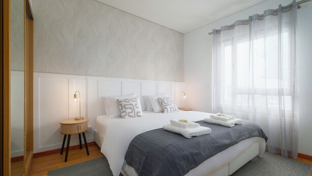 A modern bedroom with a large bed, bedside lamps, a bedside table, and a window with sheer curtains. - HALT Apartment (Ponta Delgada)