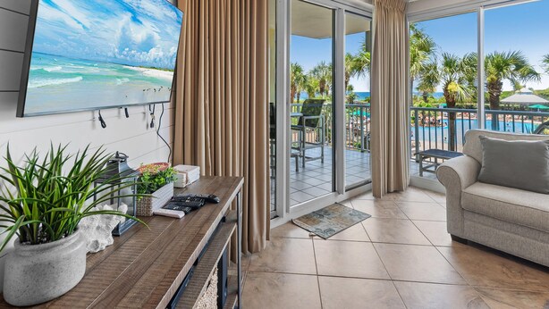 A modern living room with a large TV, a sofa, a coffee table, and a view of a pool and palm trees. - Crescent 109 (Miramar Beach)