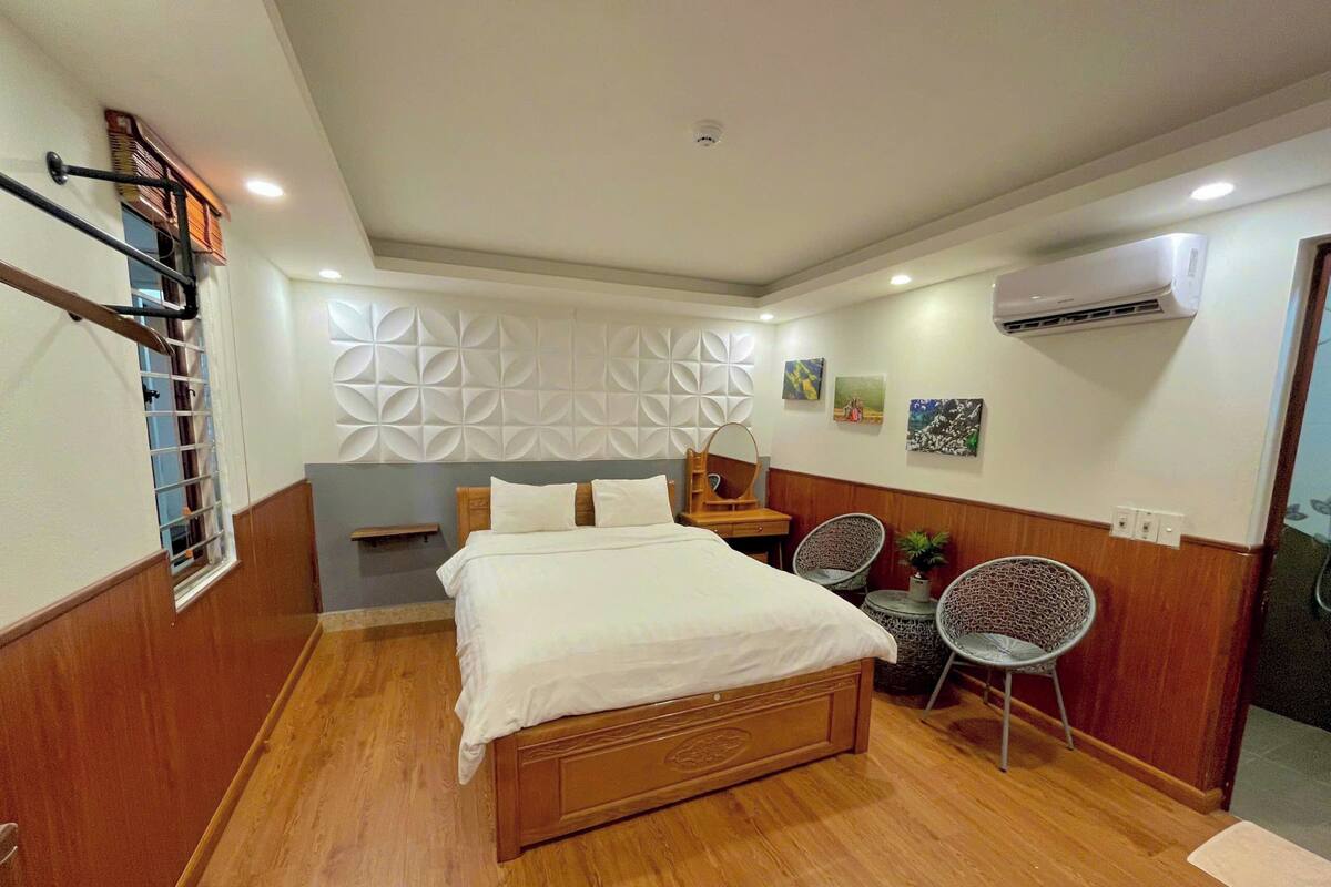 A hotel room with a wooden bed, a small desk, two chairs, a wall-mounted air conditioner, and a decorative wall panel.