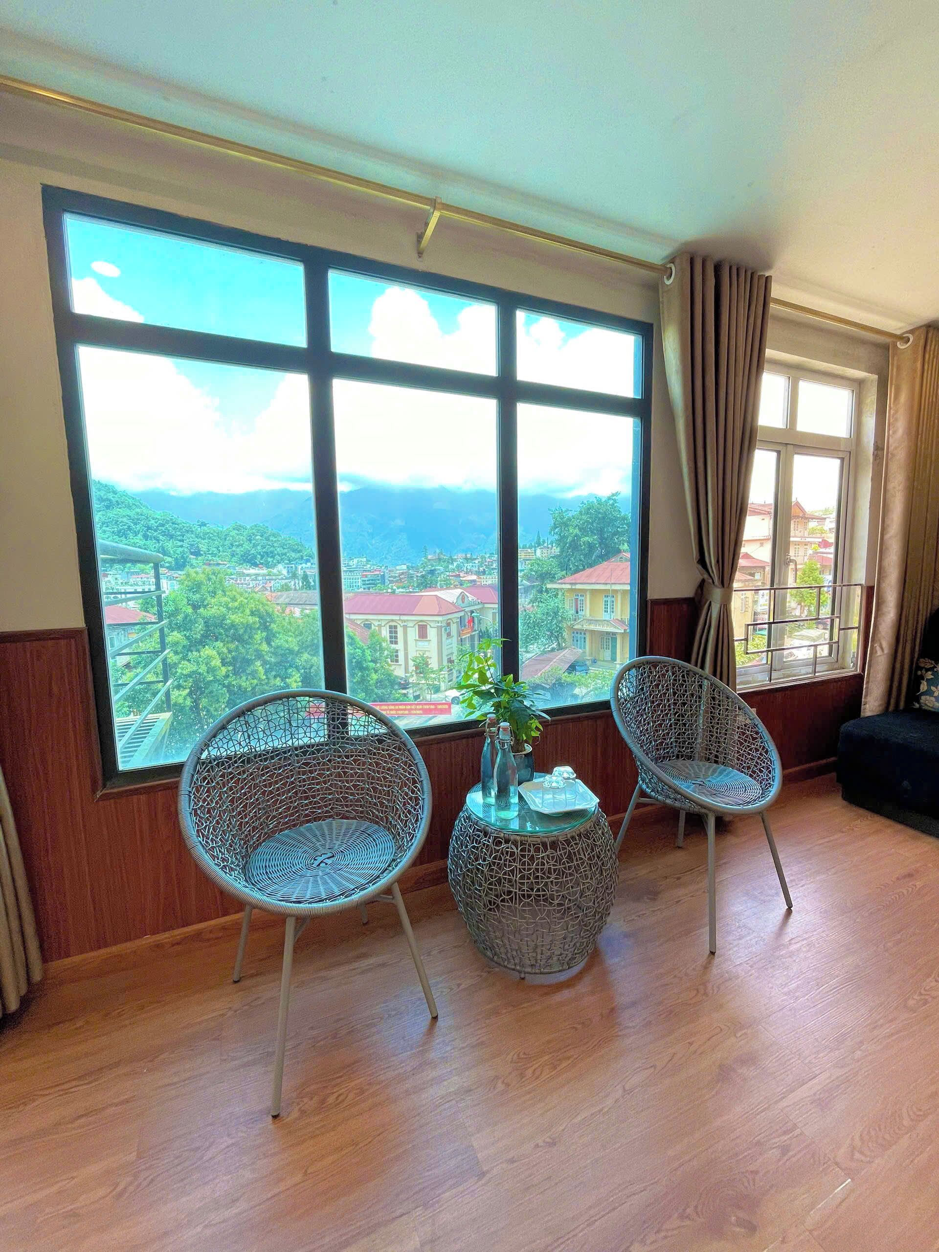 A room with large windows, two wicker chairs, a small table with a plant, and a view of a cityscape with mountains.