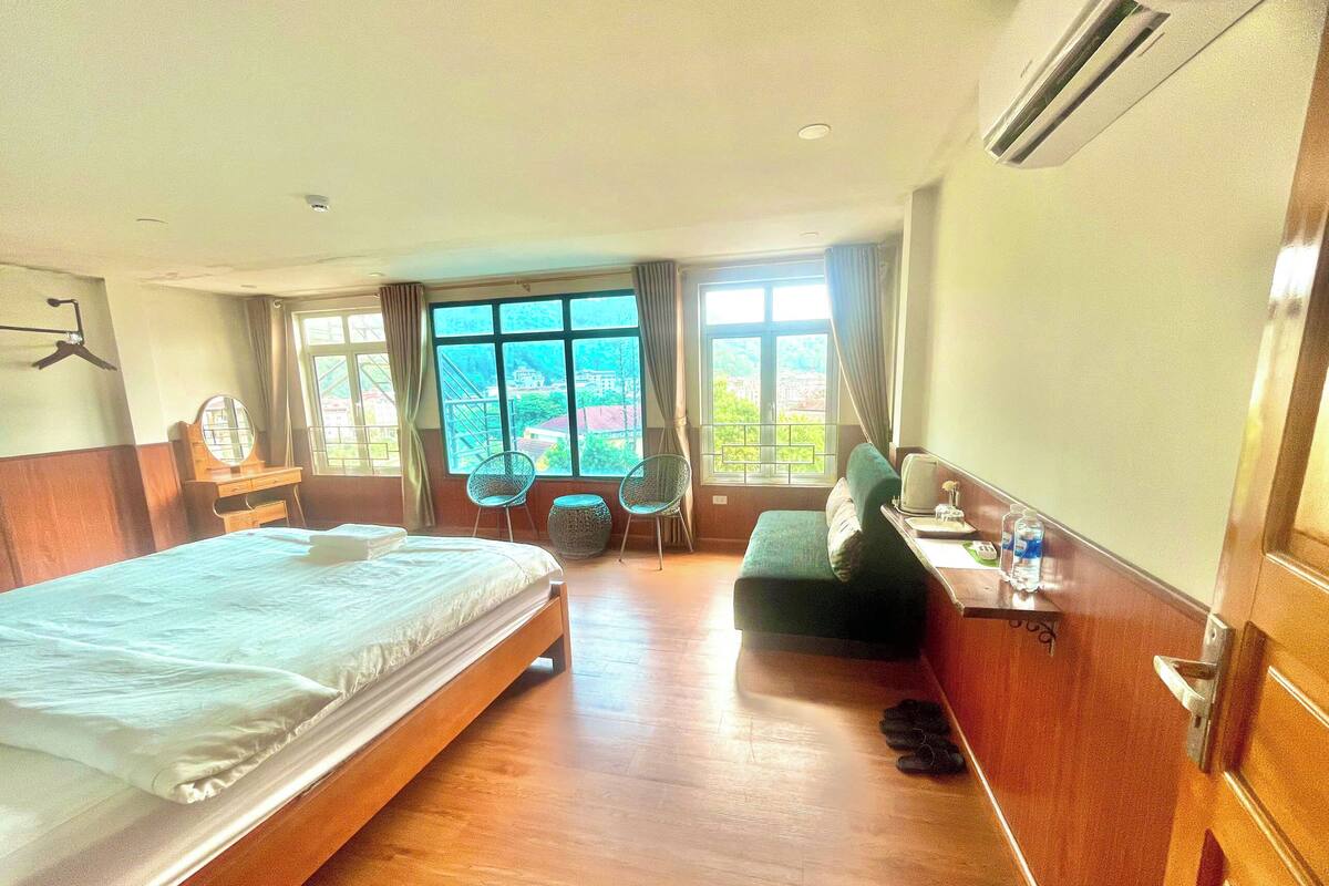 A hotel room with a large bed, a desk with a chair, a small round table, and a view of greenery through the windows.