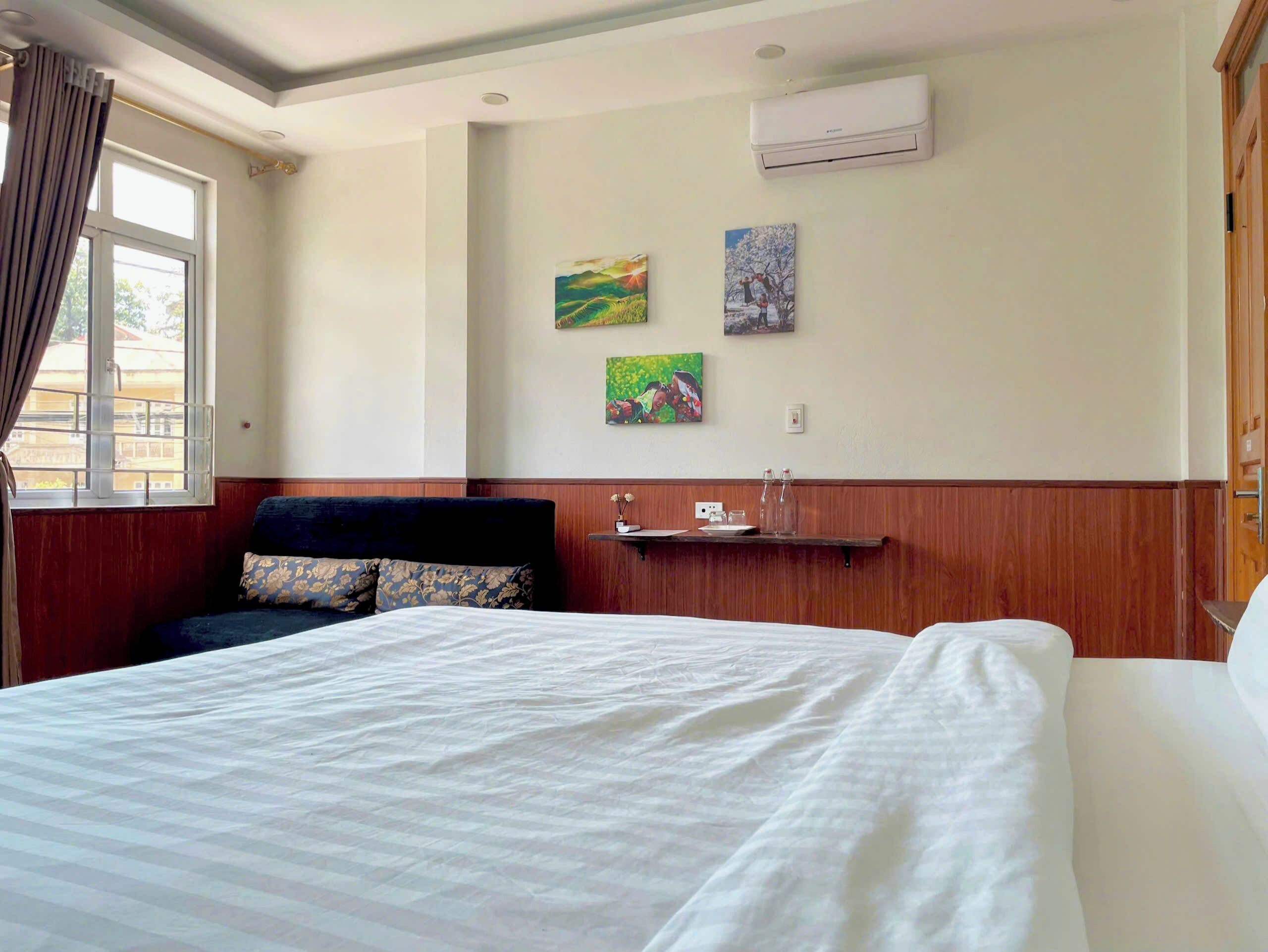 Une chambre d’hôtel avec un grand lit, un petit canapé, une étagère en bois avec quelques objets et une fenêtre avec des rideaux.