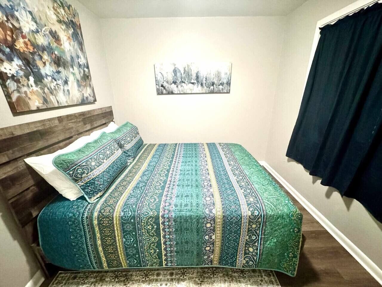 Une chambre à coucher avec un lit, une tête de lit, un tableau au mur et une fenêtre avec des rideaux.