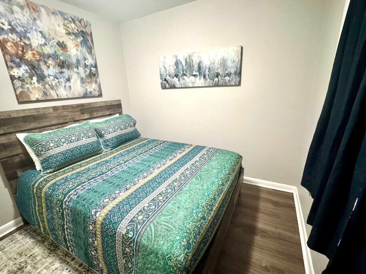 Une chambre à coucher avec un lit, deux oreillers, une tête de lit en bois, un grand tableau abstrait, un tableau représentant un paysage, plus petit, et un rideau foncé.