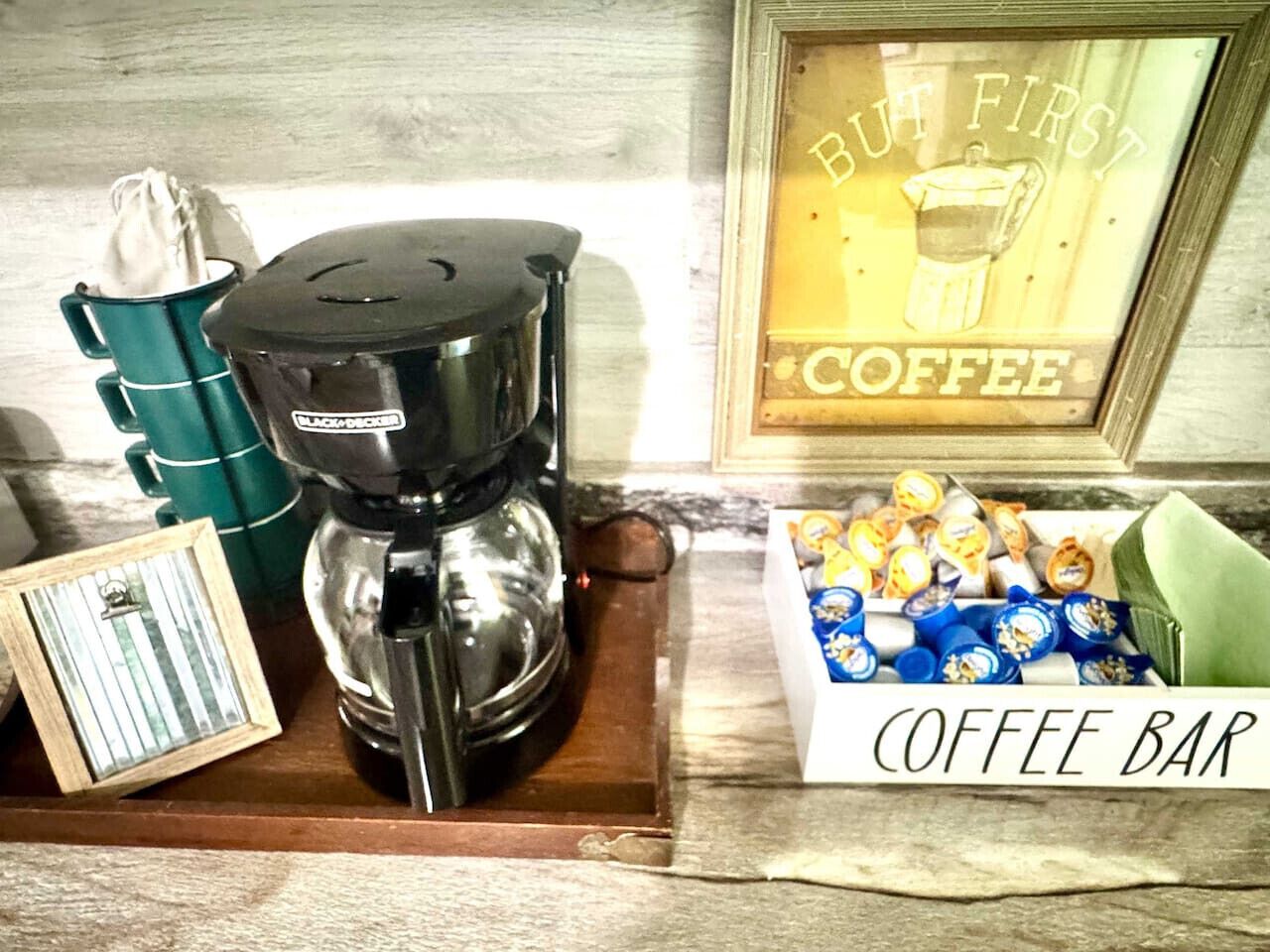 A coffee bar setup with a coffee maker, a box of coffee pods, and a framed sign that says "BUT FIRST COFFEE".