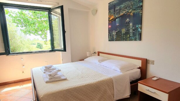 A neatly made bed with a white bedspread and pillows, a nightstand with a lamp, a window with a view of greenery, and a cityscape painting on the wall. - [Caracol 4] Apartment in the middle of the countryside with terrace (Orosei)