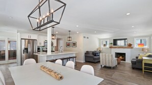 Una cocina moderna con una isla central, un comedor con una mesa blanca y una sala de estar con chimenea.