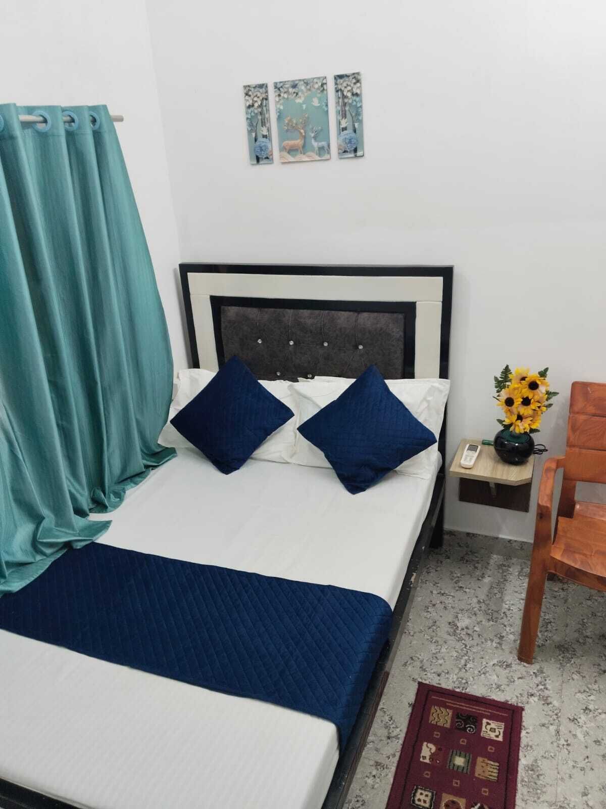 A neatly made bed with a blue quilt, a teal curtain, a bedside table with a phone and a vase of sunflowers, and a wooden chair.