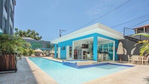 Ein moderner Poolbereich mit einem klaren, blauen Pool, einer überdachten Sitzgelegenheit und einem Gebäude mit blauer Fassade.