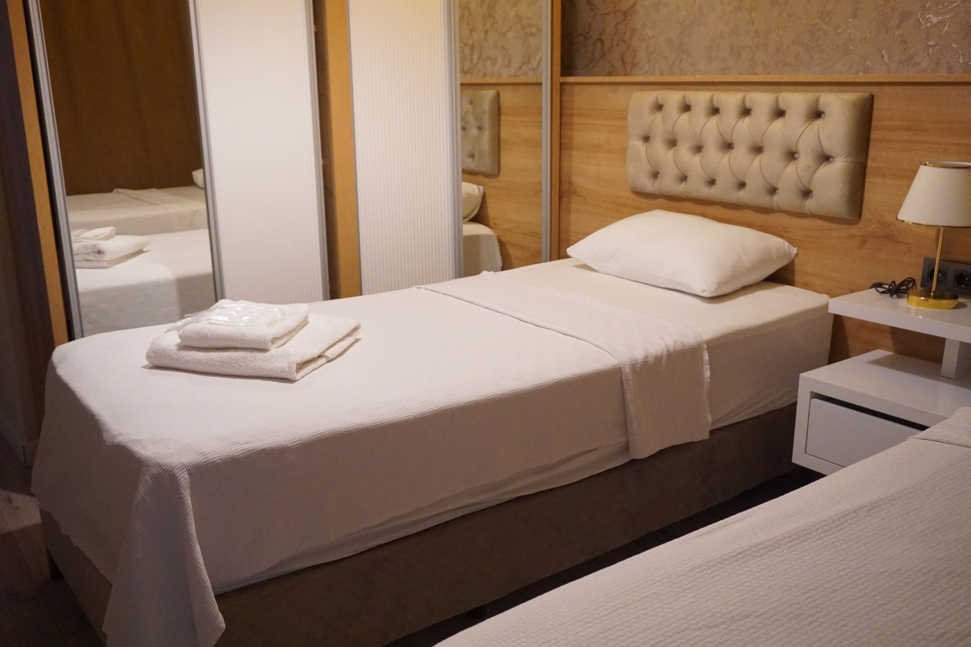 A neatly made twin bed with a white duvet and pillows, a bedside table with a lamp, and a mirror on the wall.