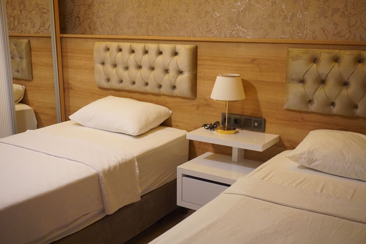 Habitación de hotel con dos camas, cada una con cabecero capitonado, una mesita de noche con lámpara y un pequeño atril.
