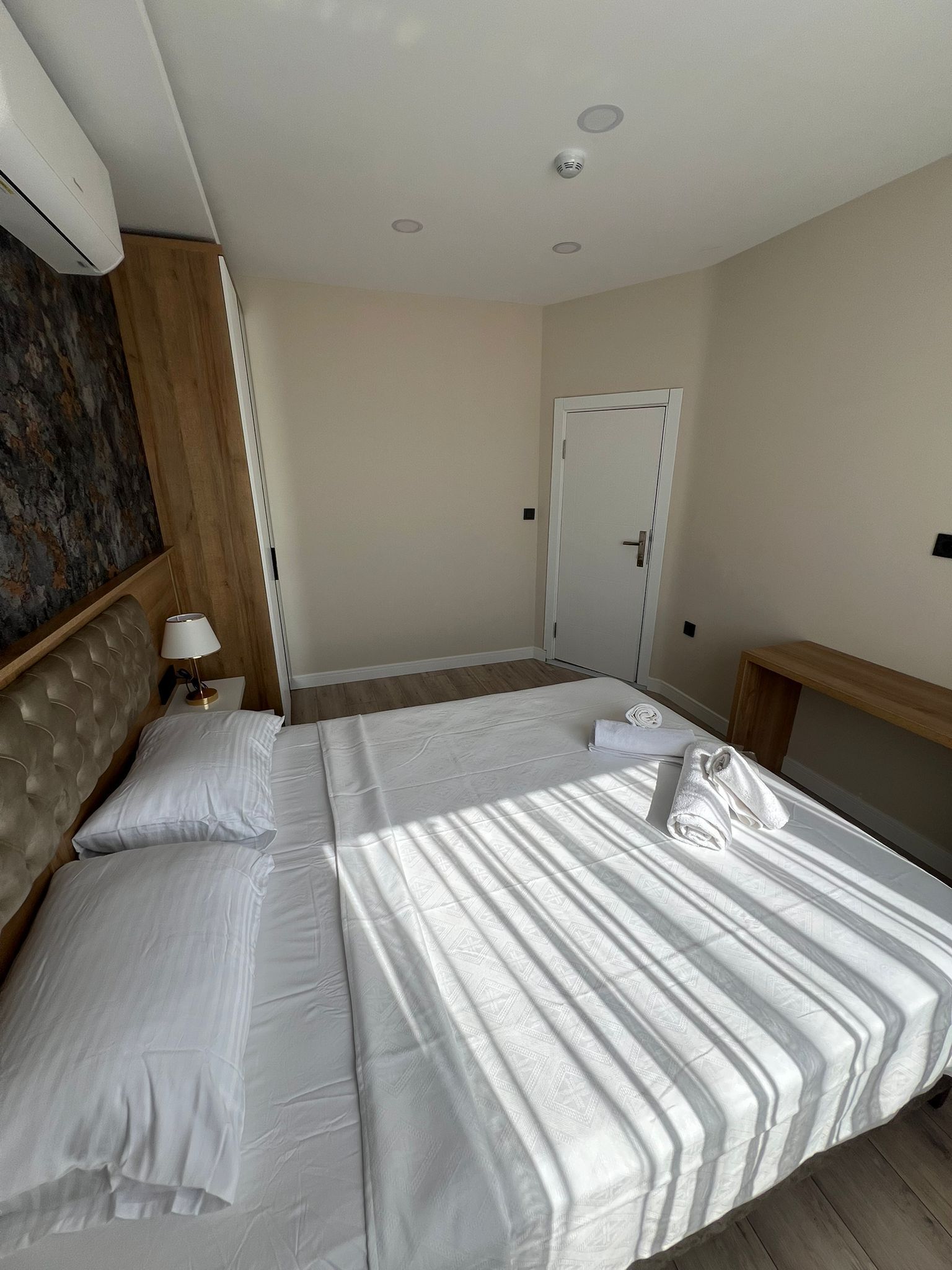 A hotel room with a bed, a headboard, a nightstand, a lamp, and a door.