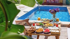Ein Esstisch mit verschiedenen Speisen und Getränken, einer Blumenvase und einem Pool im Hintergrund.