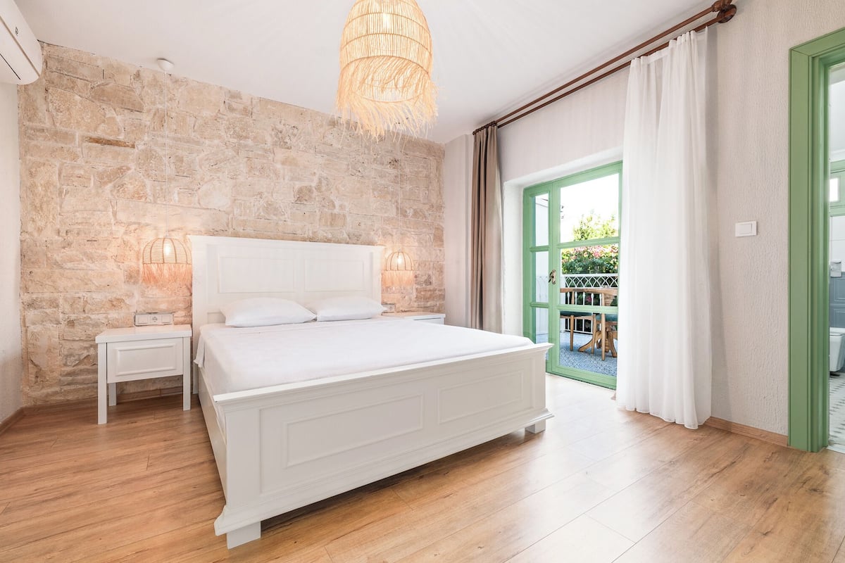A bedroom with a stone wall, a white bed, and a view of a green door leading outside.