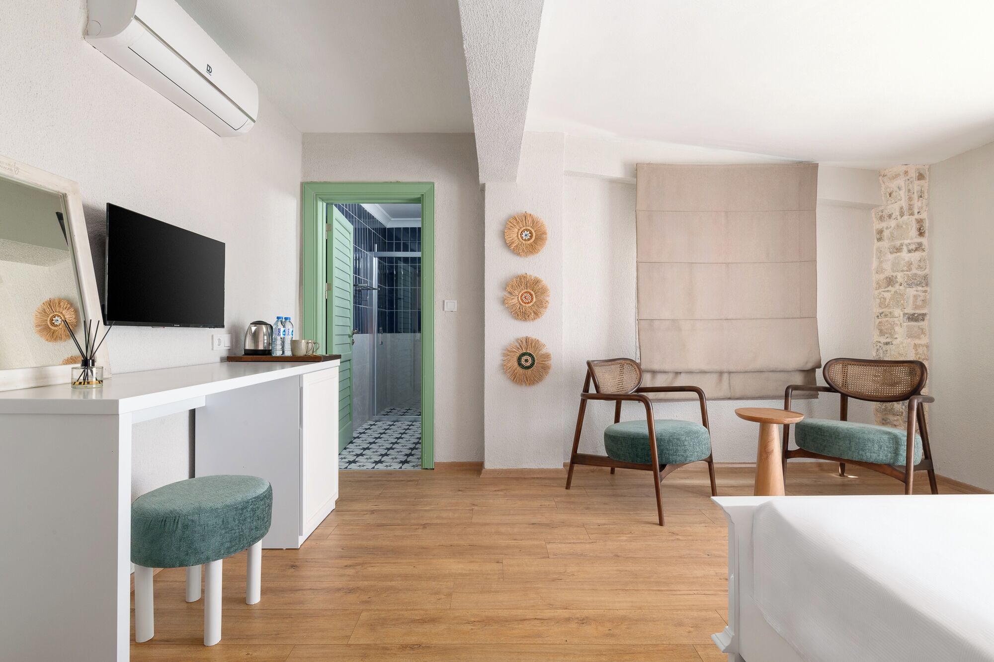 Quarto moderno com escrivaninha branca, duas cadeiras, uma mesinha e uma cama. Há uma televisão na parede, uma porta que dá para um banheiro e um enfeite de parede.