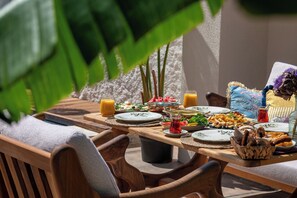 A wooden dining table set with various dishes, including fruits, bread, and drinks, in an outdoor setting with chairs and a large plant in the background. - Cerenity Alaçatı (Cesme)