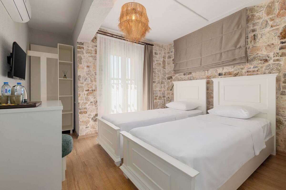 A bedroom with two white beds, a stone wall, a wooden floor, a TV on the wall, and a window with curtains.