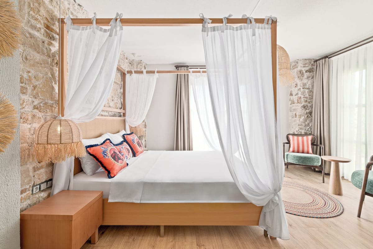 A bedroom with a four-poster bed, a bedside table, a lamp, and a view of the outdoors.