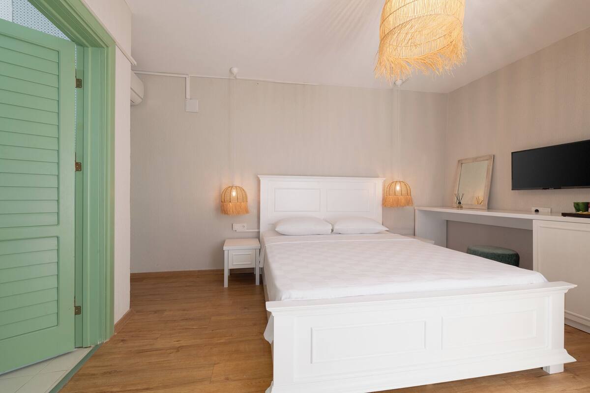 A bedroom with a white bed, a green door, a small white desk, and a mounted TV.