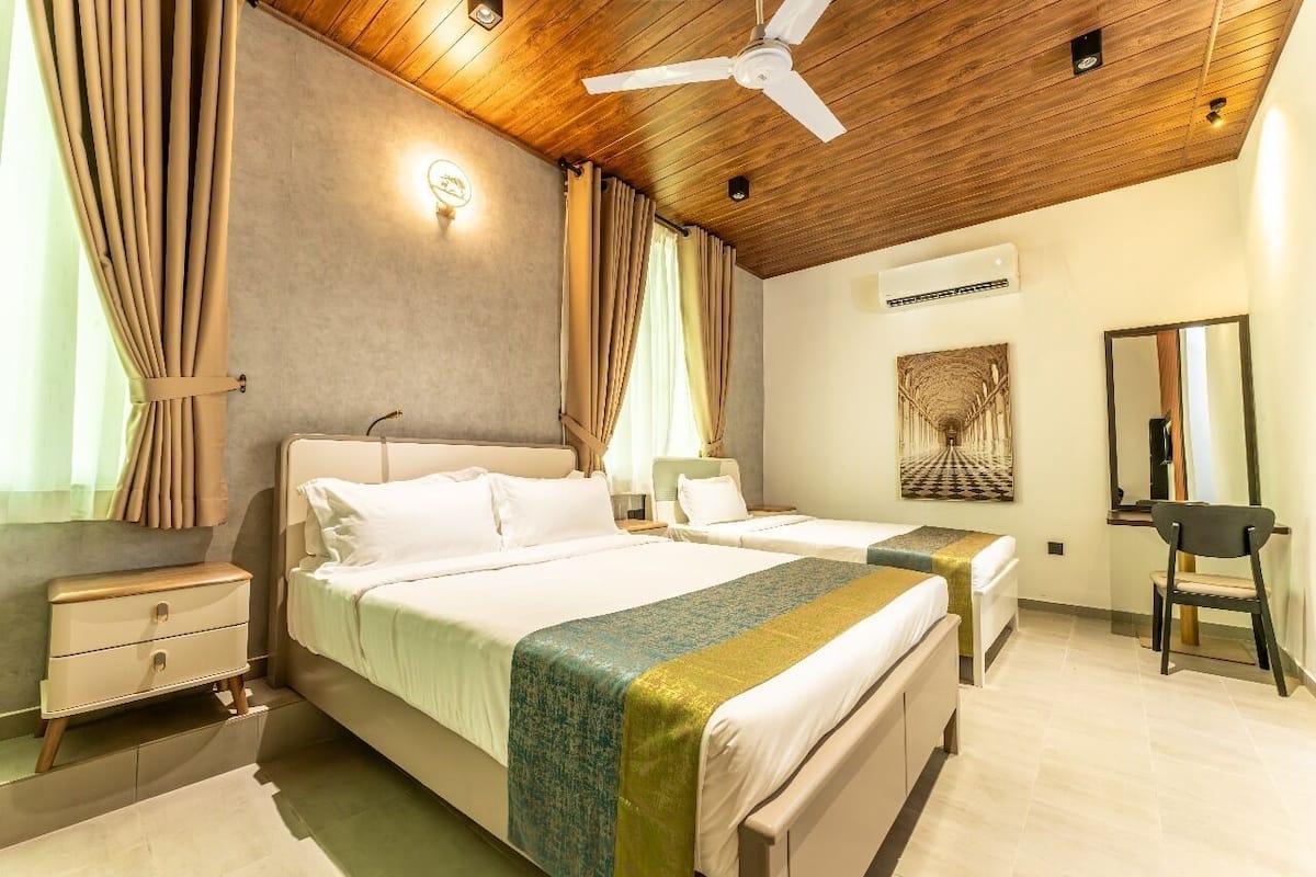 A hotel room with a wooden ceiling, two beds, a nightstand, a chair, a mirror, and a wall-mounted air conditioner.
