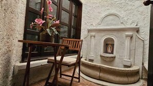 Μια αυλή με πλακόστρωτο δάπεδο, ένα ξύλινο τραπέζι και καρέκλες, ένα παράθυρο διακοσμημένο με λουλούδια και ένα τζάκι από πέτρα με μια θρησκευτική εικόνα.